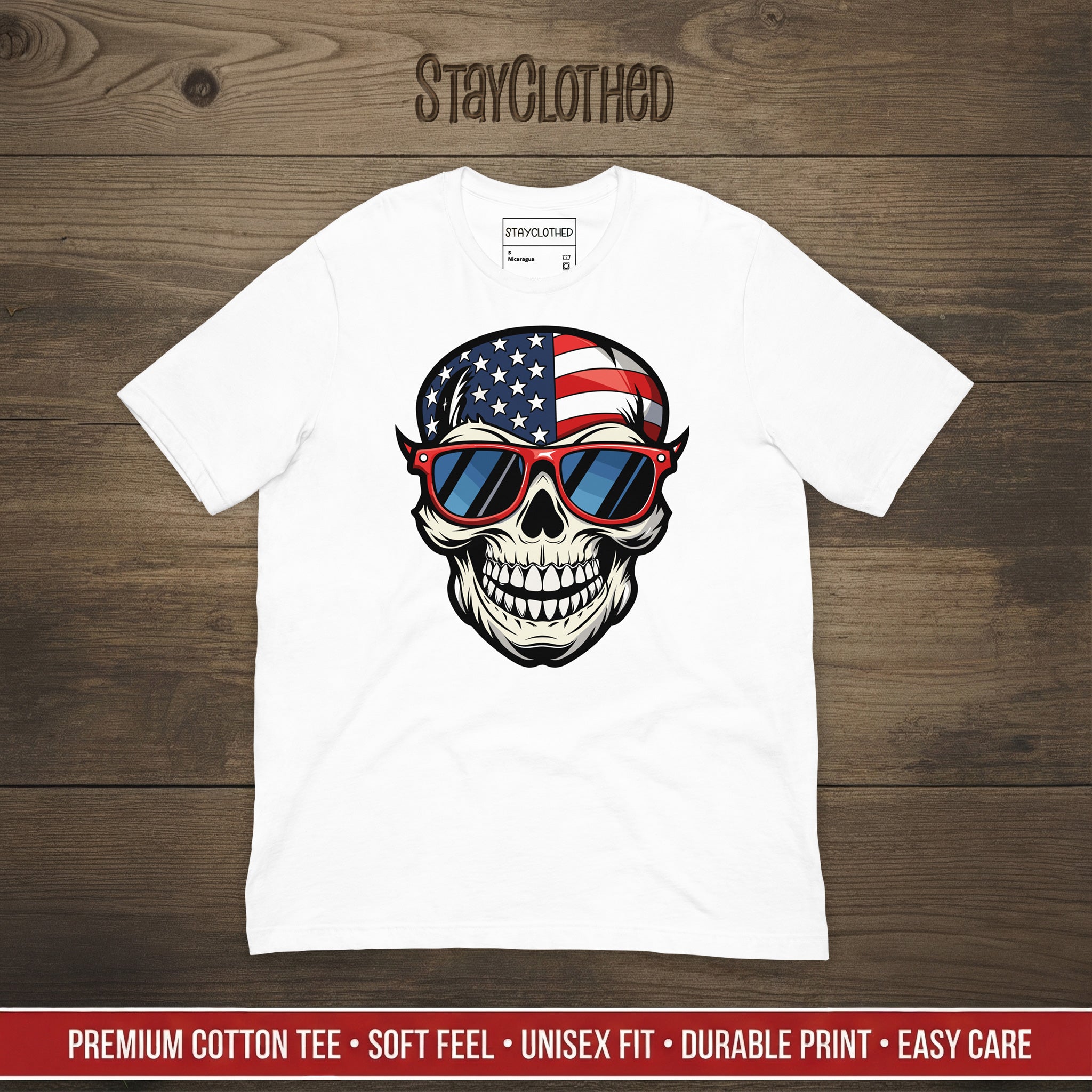 White t-shirt with a skull design wearing American flag sunglasses on a wooden surface, featuring the brand 'StayClothed'.