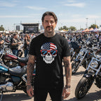 Man wearing a black t-shirt with a skull design featuring an American flag and sunglasses, standing in a motorcycle rally setting.