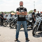 Man with tattoos wearing a black t-shirt featuring a dark skull pile design standing in front of motorcycles at a gathering