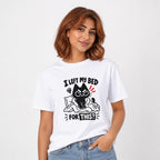 Model wearing a white T-shirt with a grumpy black cartoon cat on a rumpled bed beneath the words “I LEFT MY BED FOR THIS?”