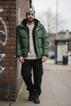 Man wearing a green puffer jacket and black pants leaning against a brick wall.