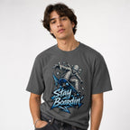 Model wearing a Pepper T-Shirt featuring a bandage-wrapped mummy riding a blue skateboard above bold “STAY BOARDIN’” lettering.