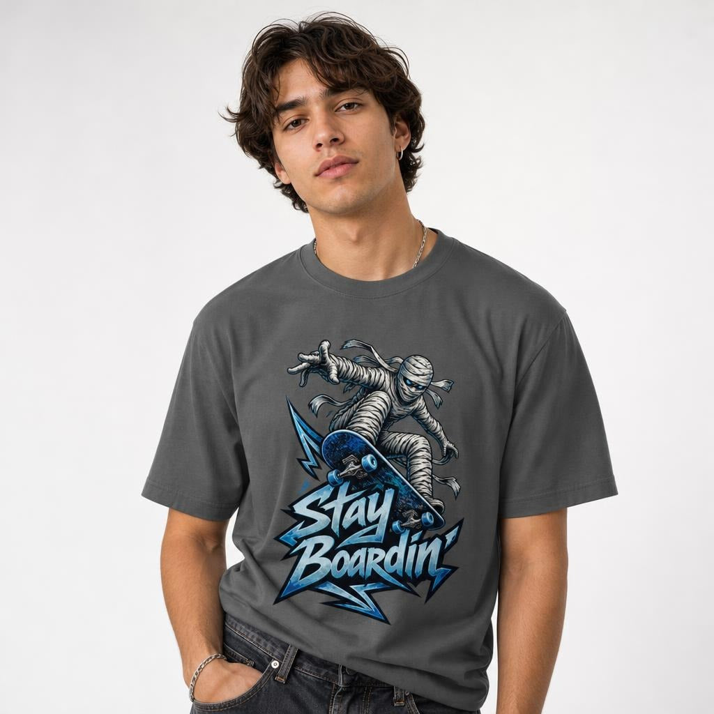Model wearing a Pepper T-Shirt featuring a bandage-wrapped mummy riding a blue skateboard above bold “STAY BOARDIN’” lettering.