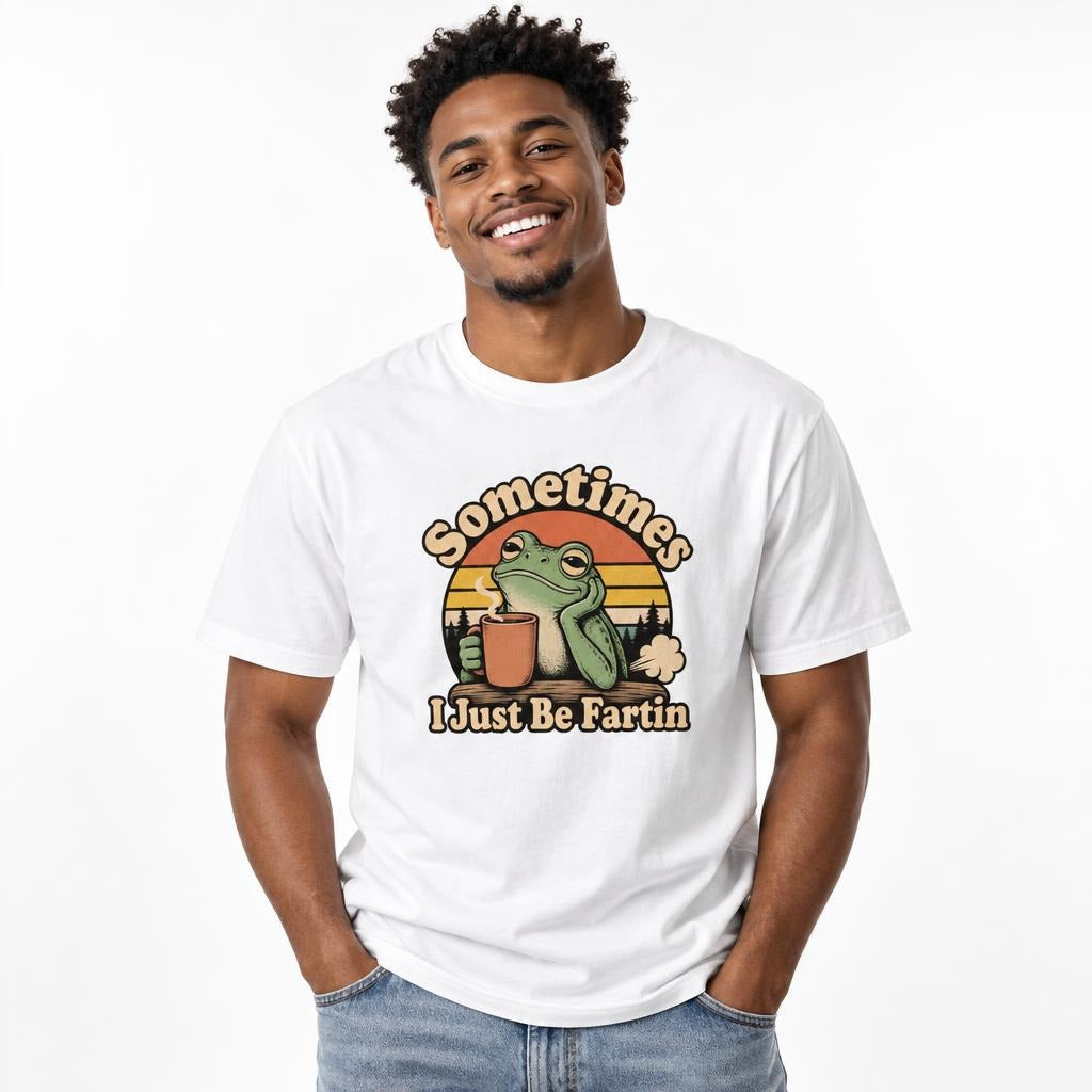 Front view of a white T-shirt with “Sometimes I Just Be Fartin” lettering, a frog holding a steaming coffee cup, and a sunset design.