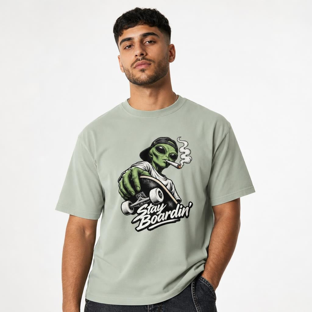 Model wearing a Bay T-shirt with a green alien in a backwards cap riding a skateboard, curling smoke and “Stay Boardin’” lettering.