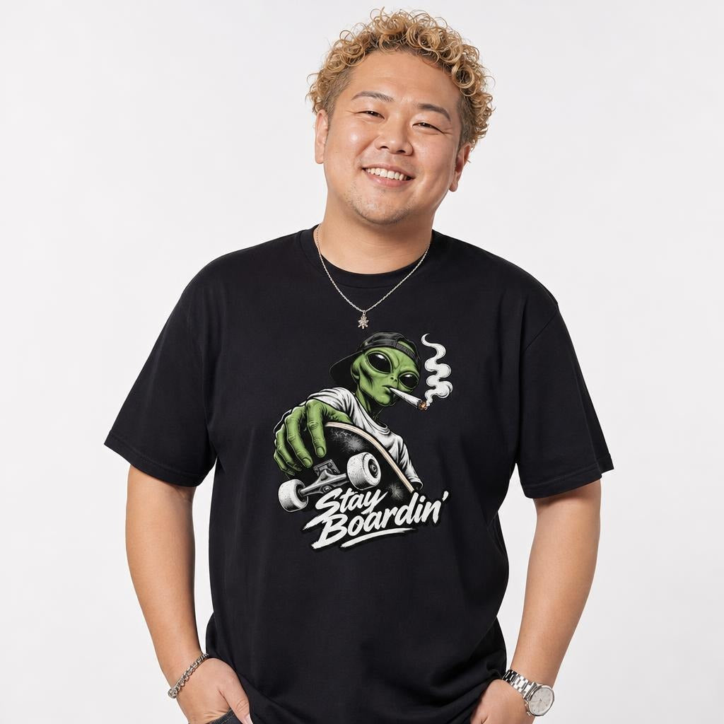 Model wearing a Black T-Shirt featuring a green alien in a backwards cap riding a skateboard, with smoke and “Stay Boardin’” lettering.