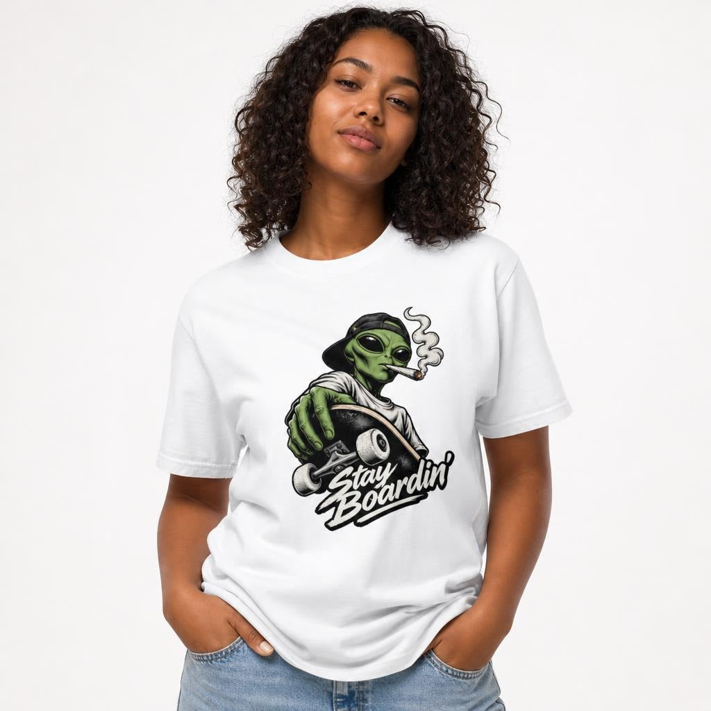 Model wearing a White T-Shirt featuring a green alien with a backwards cap riding a skateboard, smoking above “Stay Boardin’” lettering.