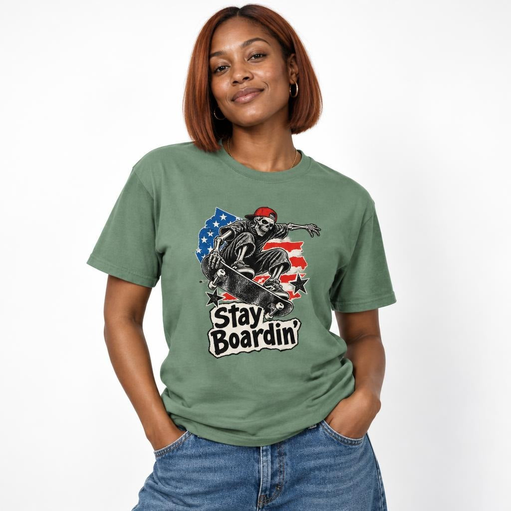 Model wearing a light green T-shirt with a front graphic of a skeleton in a red cap riding a skateboard beneath “Stay Boardin’.”