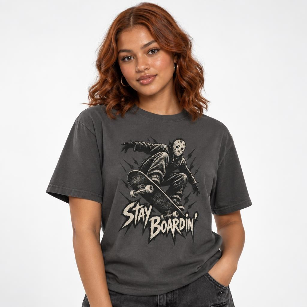 Front model view of a graphite T-shirt with a masked skater crouching on a skateboard above distressed “STAY BOARDIN’” lettering.