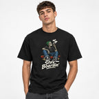 Front view of a model wearing a black T-shirt with a seated smoking skeleton on a skateboard and “Stay Boardin’” lettering.