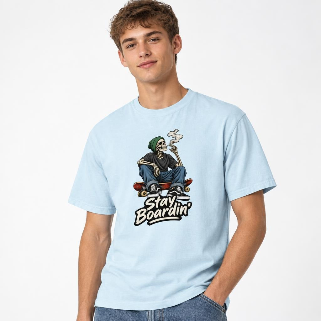Model wearing a Chambray T-shirt with a seated skeleton in a green beanie smoking on a skateboard above “Stay Boardin’” lettering.