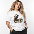 Model wearing a white T-shirt featuring a vintage gothic illustration of a raven, skull, crescent moon, candle, ivy, and Stay Spooky scroll.