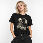 Front view of a model wearing a Black T-Shirt with a seated skeleton, black cat, crescent moon, and “Stay Spooky” banner.