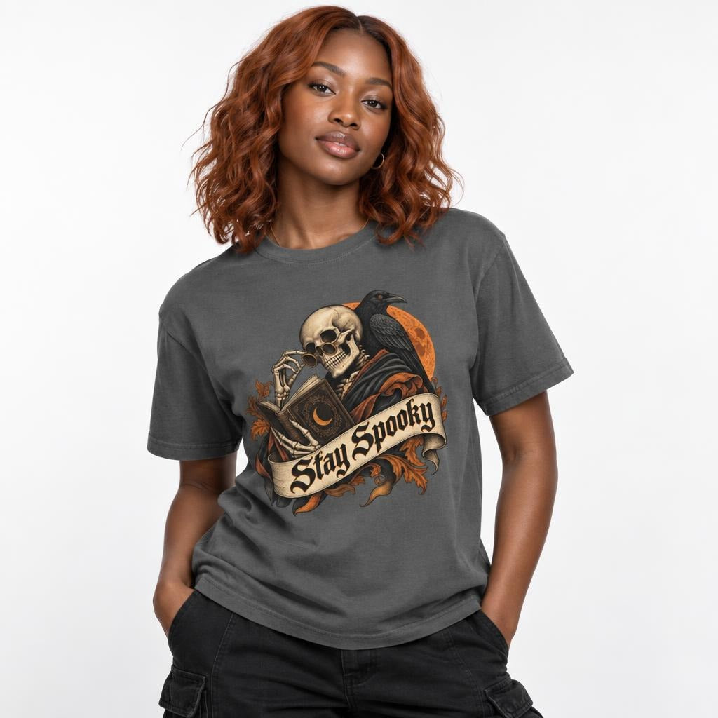 Model wearing a Pepper T-shirt with a gothic Skull, Raven, and Moon illustration, a skeleton reading beneath a “Stay Spooky” banner.
