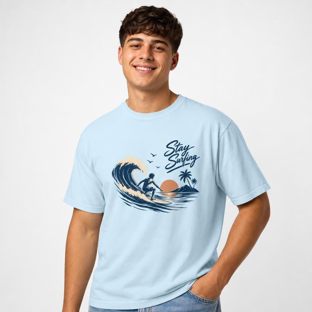 Front view of a Chambray T-shirt featuring “Stay Surfing,” a surfer riding a wave, palm trees, sunset, shoreline and birds.