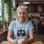 Woman sitting at a cafe holding a coffee cup, wearing a t-shirt with a cat graphic and text.