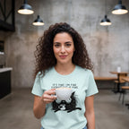 Woman wearing a light blue t-shirt with a graphic and text, holding a white mug in a casual setting.