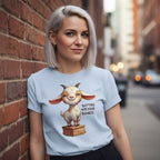 Woman wearing a light blue t-shirt with a cartoon goat graphic and text, standing against a brick wall.