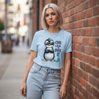 Woman wearing a light blue t-shirt with a penguin graphic and text, leaning against a brick wall.