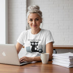 Woman wearing a white t-shirt with a graphic design, sitting at a desk with a laptop and coffee.
