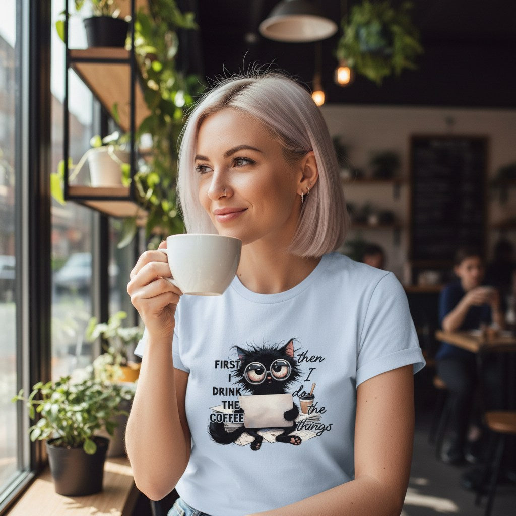 Woman in a cafe holding a cup, wearing a t-shirt with a cat graphic and text.