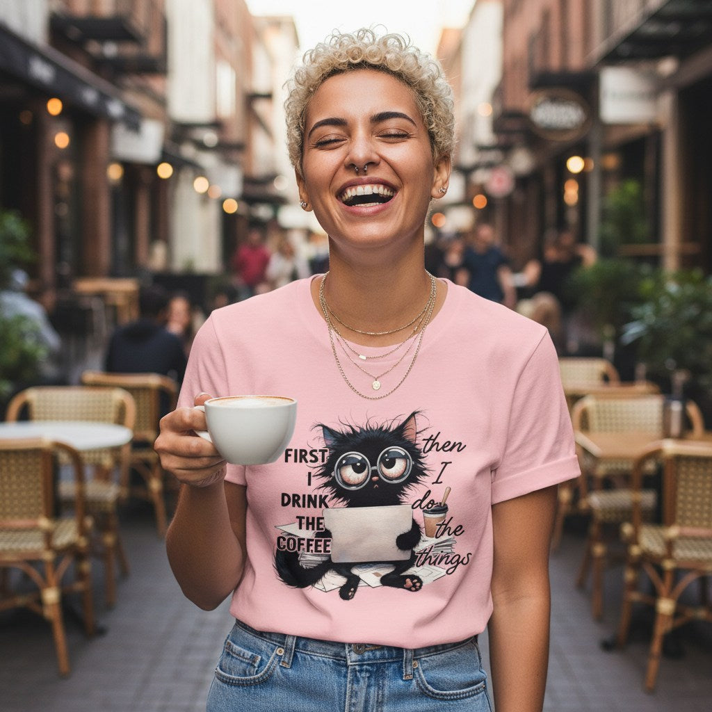 Woman holding a coffee cup and wearing a pink t-shirt with a cat graphic in an outdoor cafe setting.