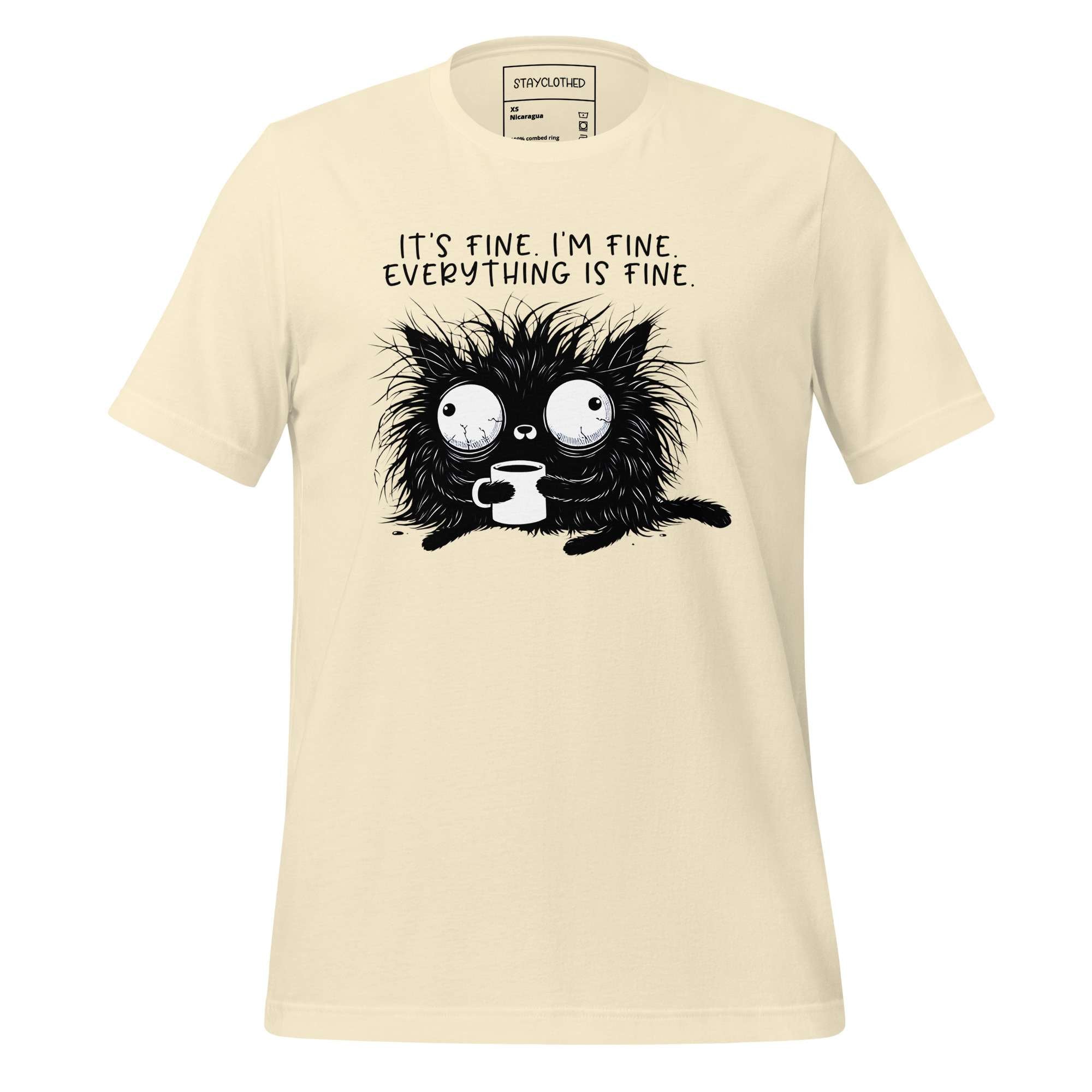 Beige t-shirt with a black cat graphic and text on a wooden surface, featuring the brand 'StayClothed'.