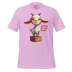 Pink t-shirt with a cartoon goat graphic and text on a wooden surface, featuring the brand 'StayClothed'.