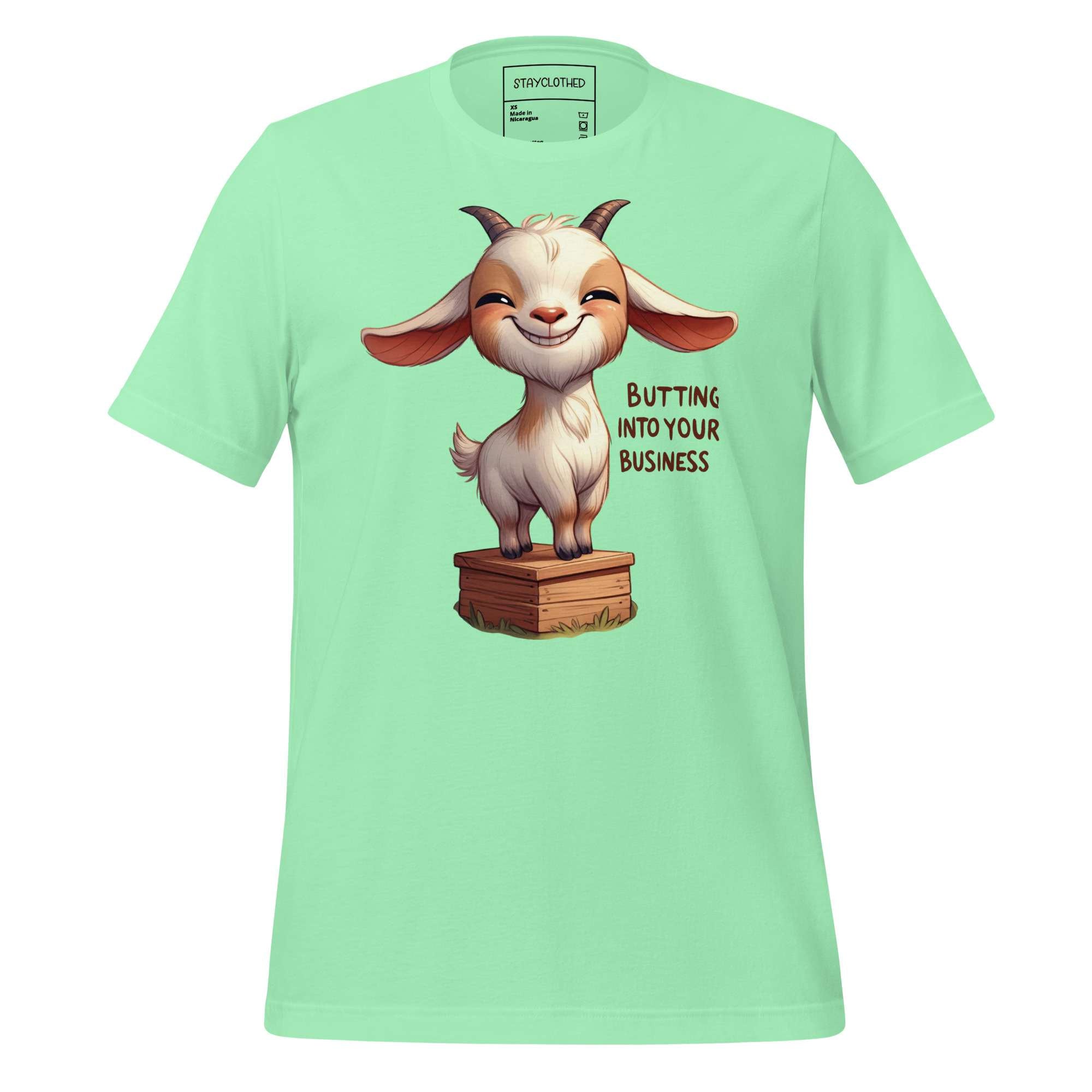 Light green t-shirt with a cartoon goat graphic and text on a wooden surface, featuring the brand 'StayClothed'.