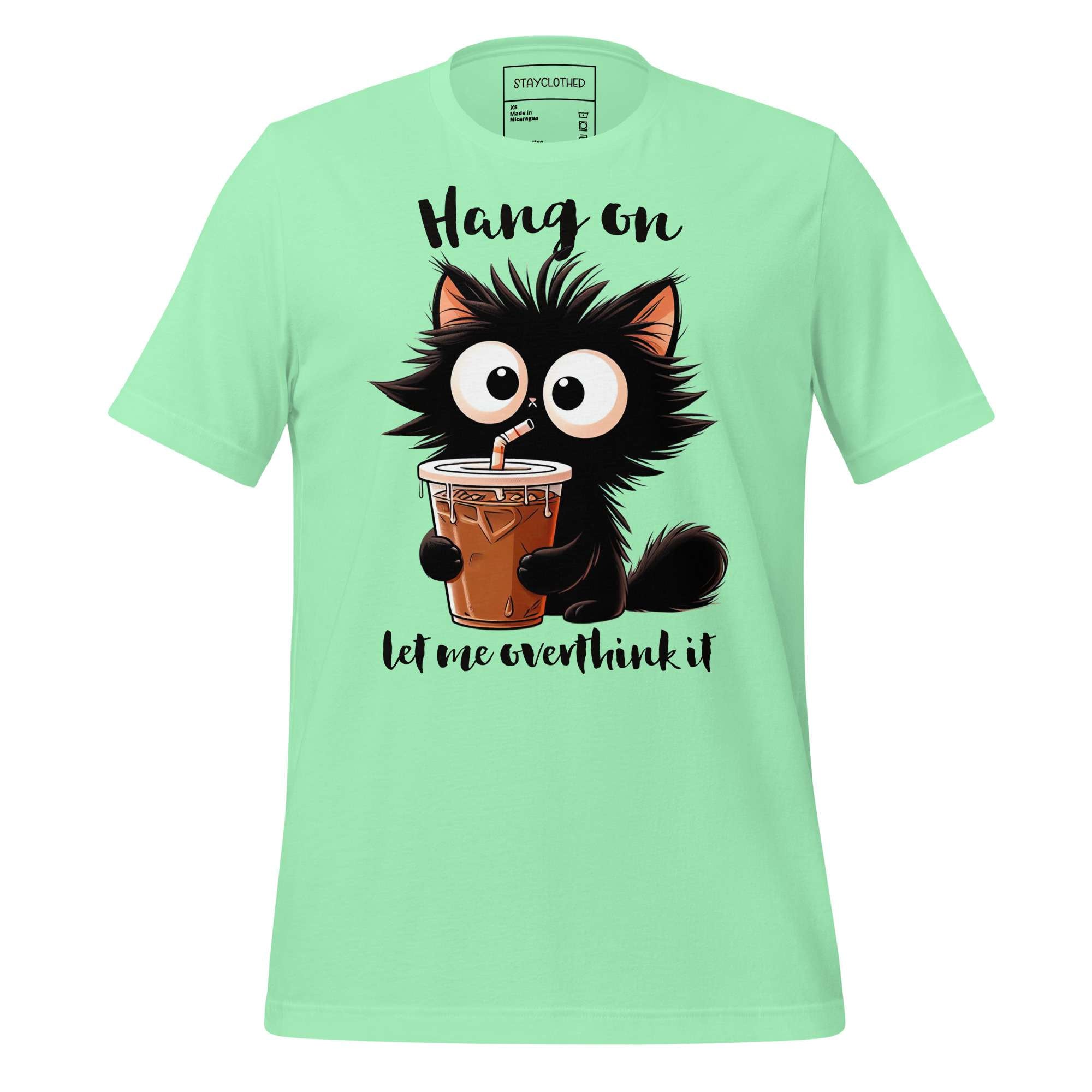 Light green t-shirt with a cartoon cat holding a drink on a wooden surface, featuring the brand 'StayClothed'.