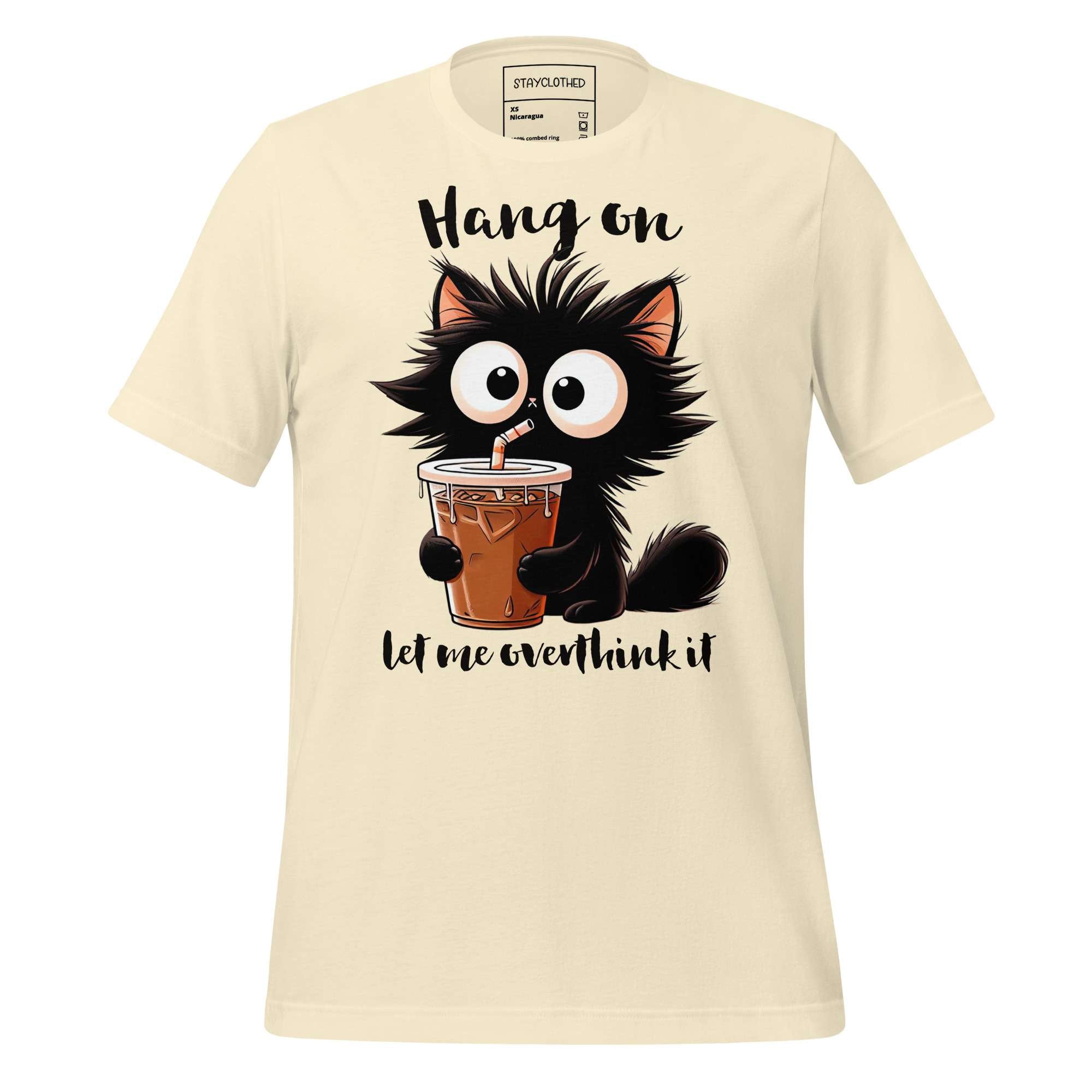 Beige t-shirt with a cartoon cat holding a drink on a wooden surface, featuring the brand 'StayClothed'.