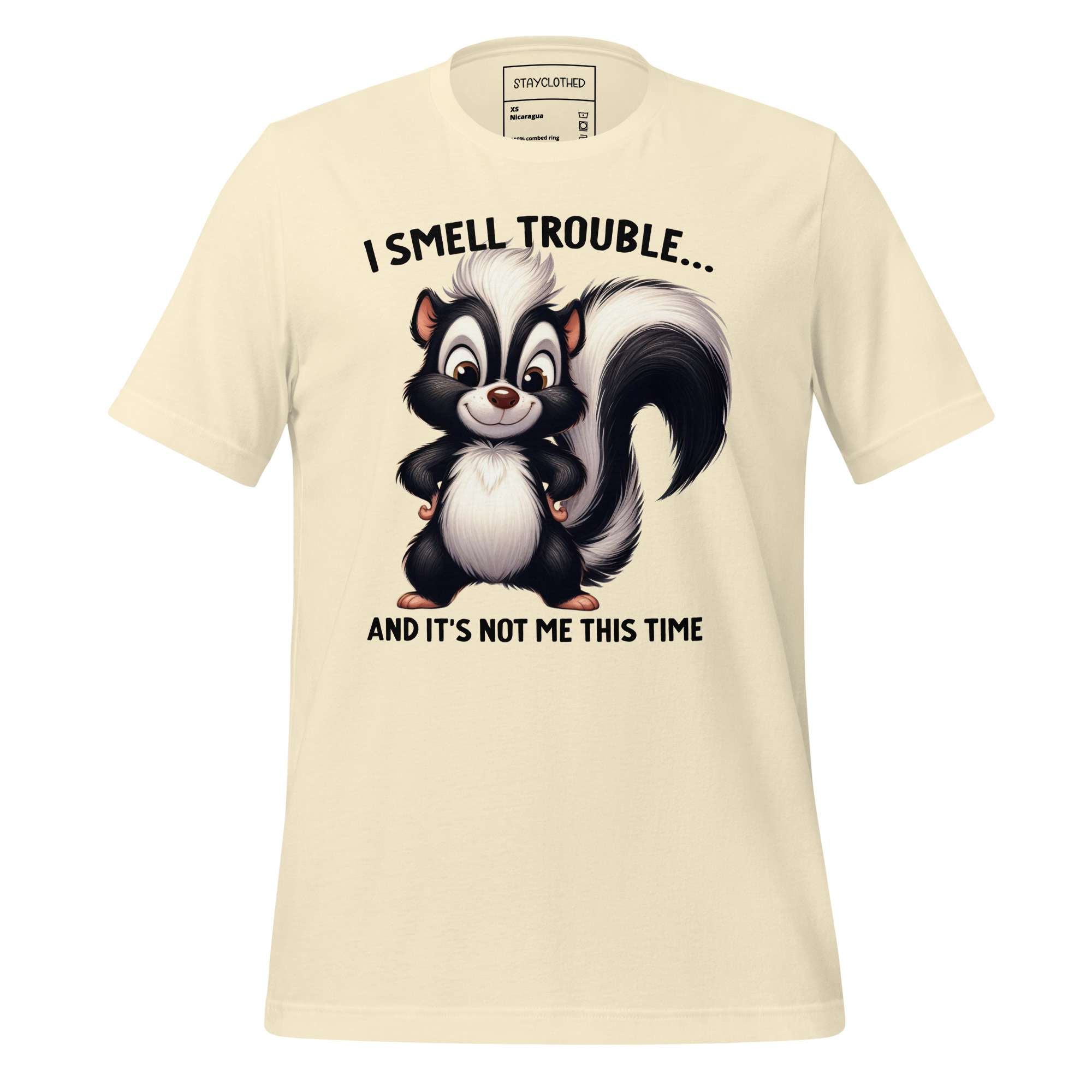 Beige t-shirt with cartoon skunk graphic and text on a wooden surface, featuring 'StayClothed' brand.