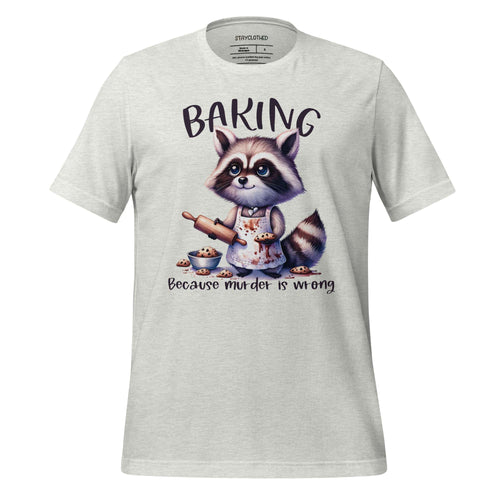 White t-shirt with a raccoon graphic and 'Baking Because Murder is Wrong' text on a wooden surface, featuring the brand 'StayClothed'.