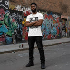 Man standing in front of a graffiti-covered wall wearing a white t-shirt featuring a dark skull pile design.