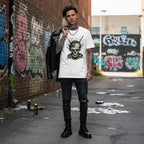 Person wearing a white t-shirt with a grimdark horror skull design, black leather jacket, and ripped jeans standing in an urban alley with graffiti.